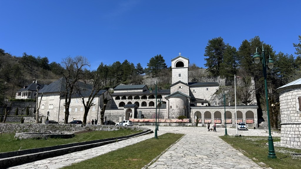 A scenic view of a stone monastery complex set against a blue sky, surrounded by trees. The structure features a bell tower and arched walkways, with a pathway leading to it.