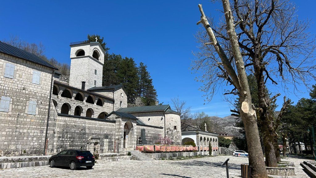 A view of a stone building with a tower set against a clear blue sky, flanked by bare trees and a parked car in the foreground.