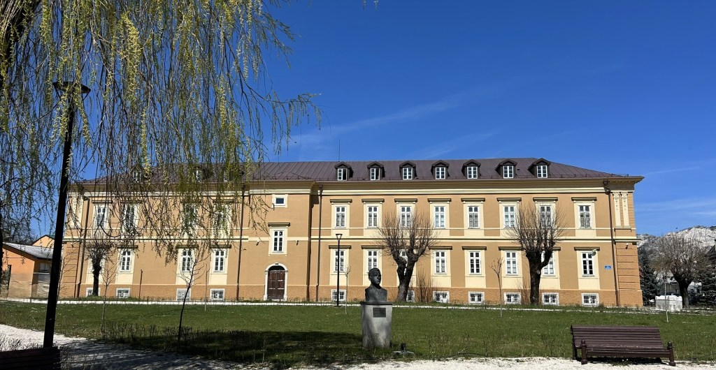 A large yellow building with a pitched roof and multiple windows, set against a clear blue sky. In the foreground, a bust statue and manicured grass area with trees are visible.