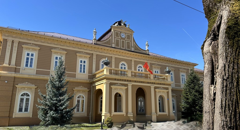 A historic building with a decorative façade, featuring large windows and a clock at the top. The entrance has a balcony with a red flag, framed by trees against a clear blue sky.