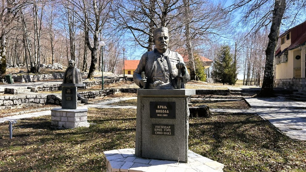 A bronze statue of King Nikola I, 1841-1921, surrounded by trees in a park setting, with another bust visible in the background.