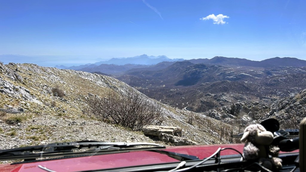 View from inside a vehicle overlooking a mountainous landscape with rocky terrain and distant peaks under a clear blue sky.