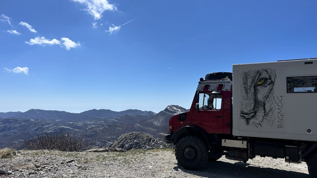 A red overland vehicle with a lion illustration on its side, parked on a rocky hilltop overlooking a mountainous landscape under a blue sky with scattered clouds.