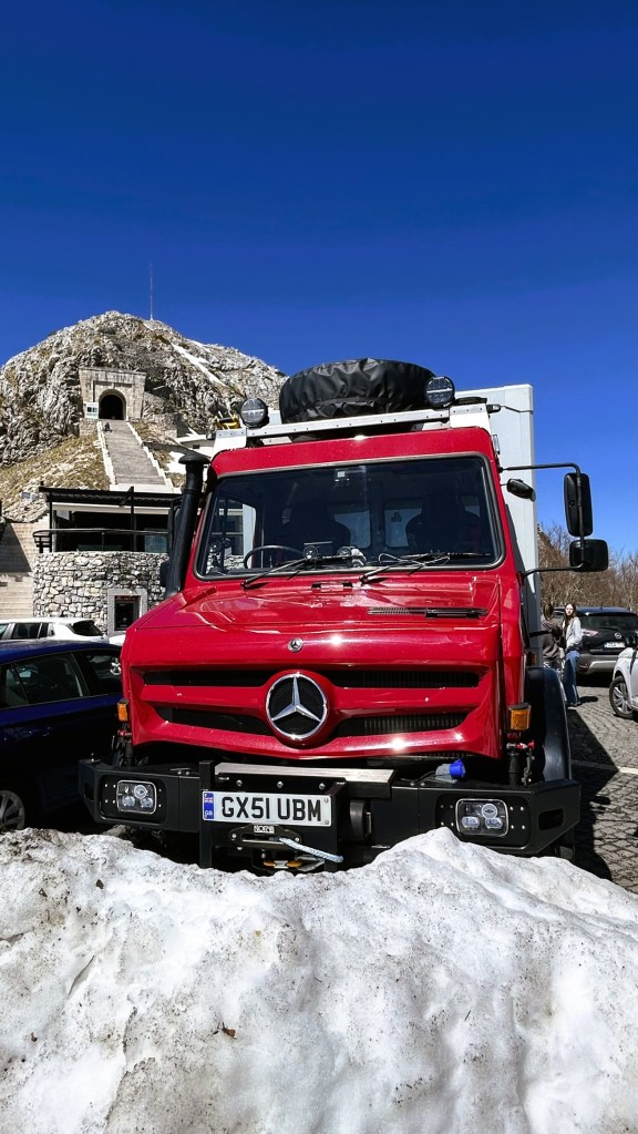 A red Mercedes truck parked on snow in front of a mountain with a clear blue sky.