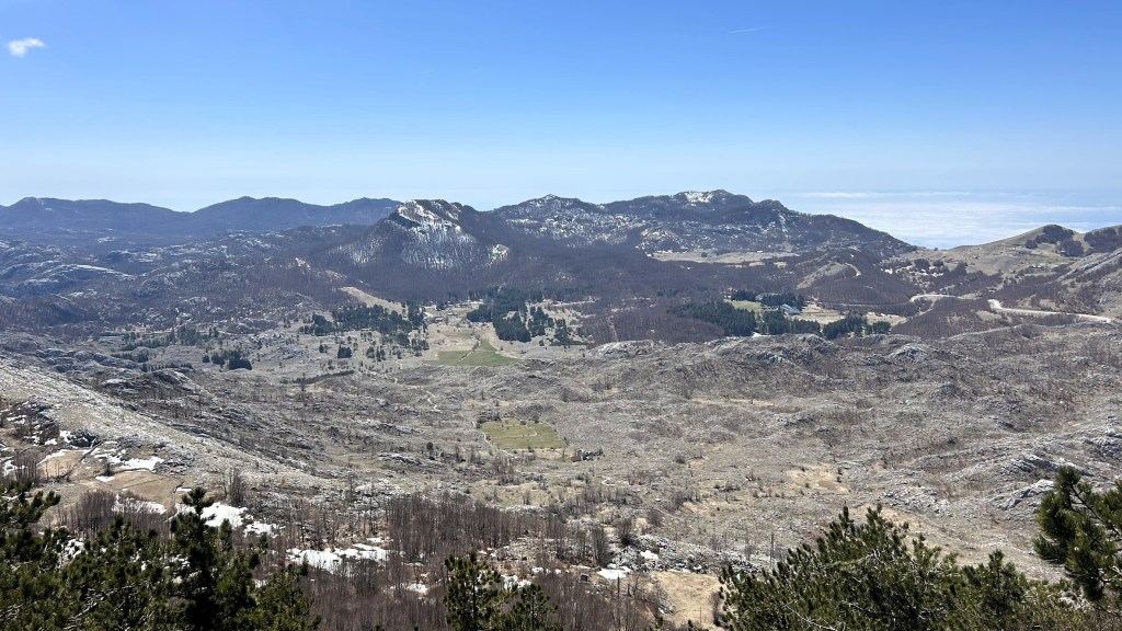 A panoramic view of a mountainous landscape with a mix of rocky terrain and patches of greenery. The sky is clear and blue, showcasing distant mountains and valleys.