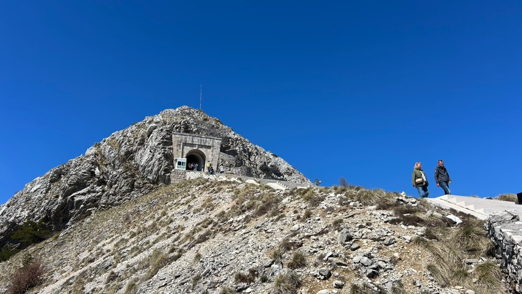 Two people walking on a rocky path towards a mountain entrance under a clear blue sky.