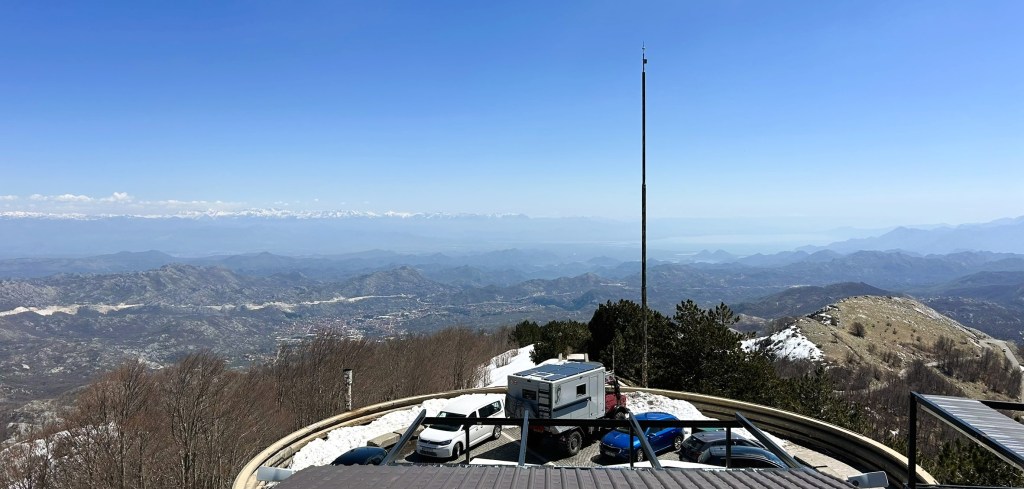A panoramic view from a mountain peak, showing a vast landscape of hills and valleys below, with a clear blue sky and some snow-capped mountains in the distance. In the foreground, there is a small building and parked cars.