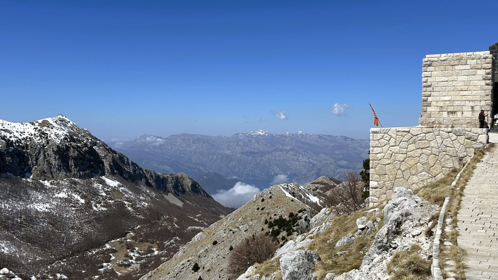A panoramic view of mountain ranges with snowy peaks and a clear blue sky, featuring a stone structure on the right and a winding path leading down the slope.