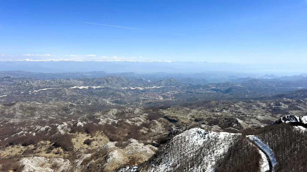 Panoramic view of snow-capped mountains and rolling hills under a clear blue sky, with a small town visible in the distance.