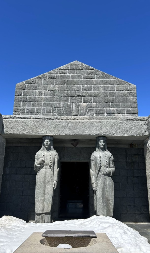 A stone structure featuring two large statues of figures standing by the entrance, with a clear blue sky above and some snow in the foreground.