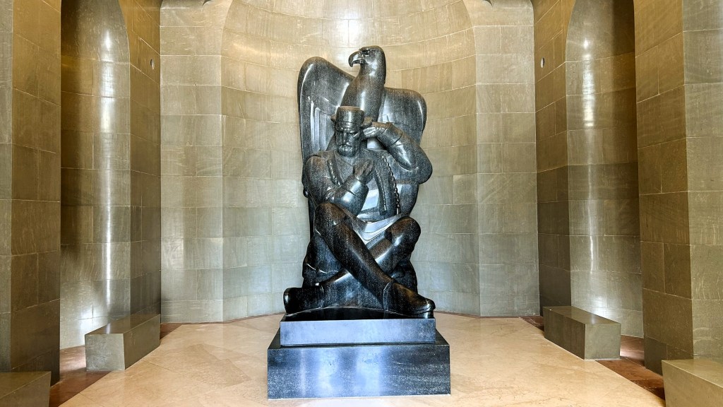A large black statue of a seated figure wearing a military-style uniform, with an eagle perched behind them, set in a minimalist stone hall.