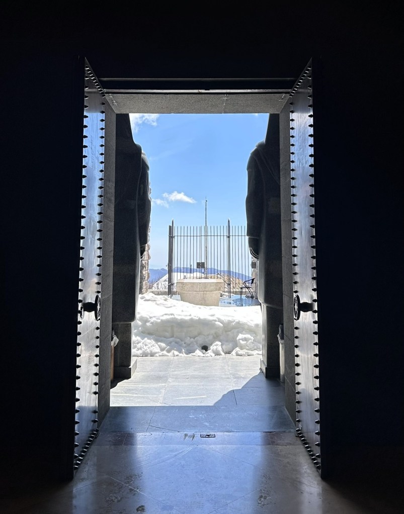 View through an open door framed by large stone statues, revealing a snow-covered landscape and blue sky beyond the doorway.