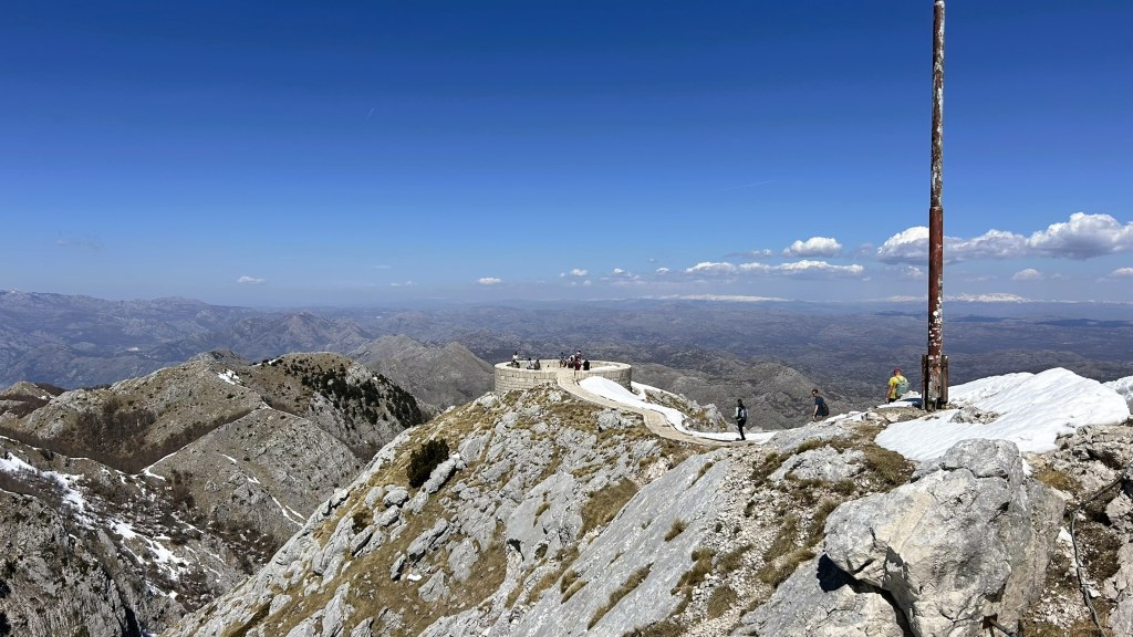 Scenic view from a mountain peak with a pathway and visitors, surrounded by rocky terrain and distant mountains under a clear blue sky.