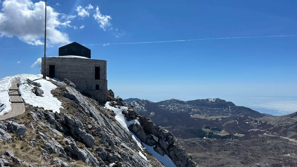A stone building with a flat roof situated on a rocky mountain summit, surrounded by patches of snow, against a clear blue sky.