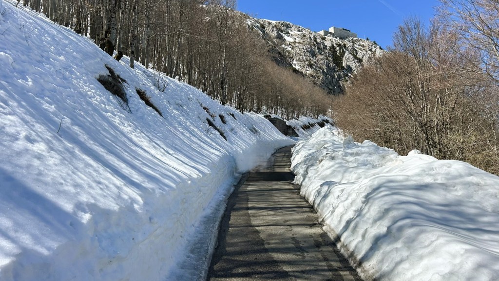 A snow-covered road flanked by tall snowbanks and bare trees, with rocky hills visible in the background under a clear blue sky.