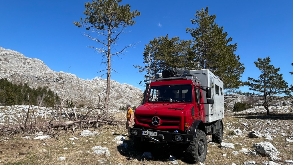 A red Mercedes off-road vehicle parked in a rocky landscape, surrounded by pine trees and mountains under a clear blue sky.