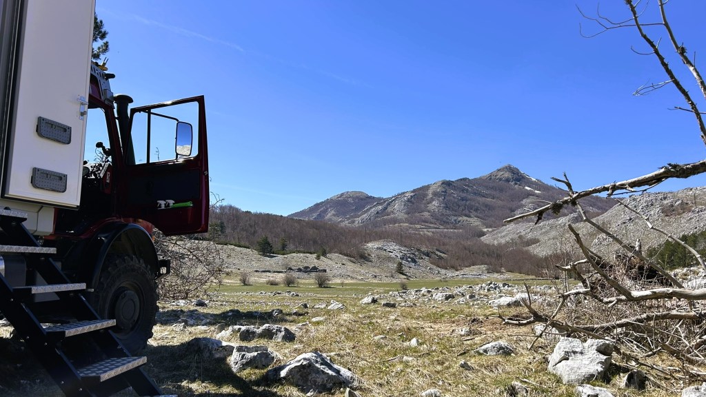 A rugged landscape with mountains in the background, featuring an open door of a truck and steps leading up to it, surrounded by rocky terrain and sparse vegetation under a clear blue sky.