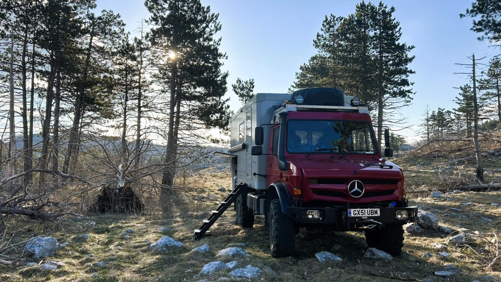A red Mercedes off-road vehicle parked in a forested area with tall trees and rocky ground, sunlight filtering through the branches.