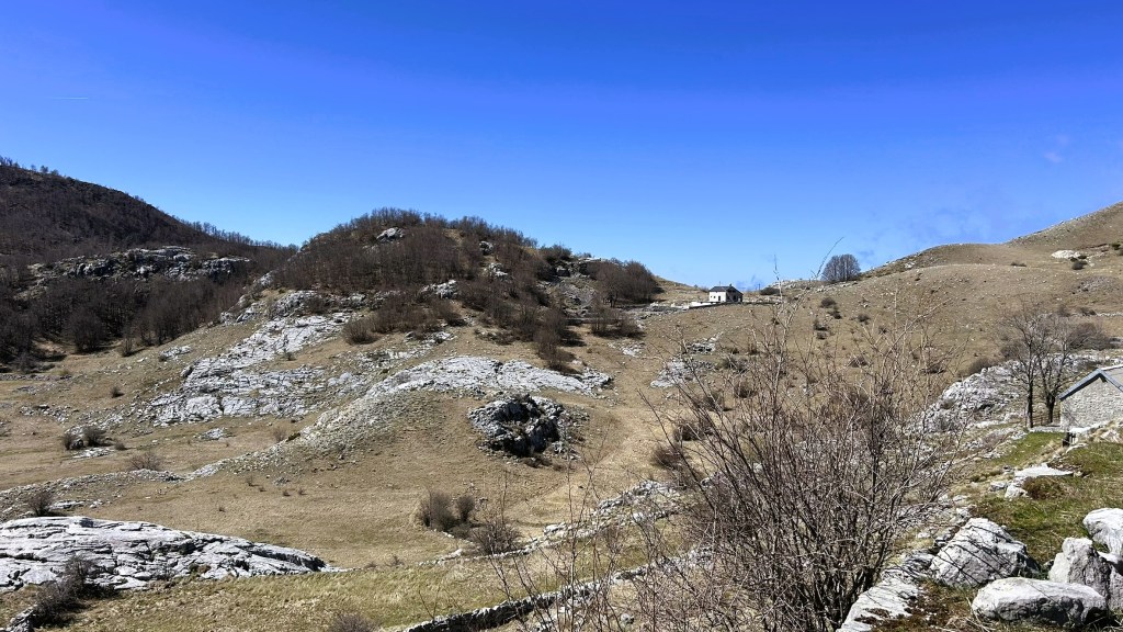 A scenic landscape featuring rolling hills and rocky outcrops under a clear blue sky, with a small house in the distance and sparse vegetation.
