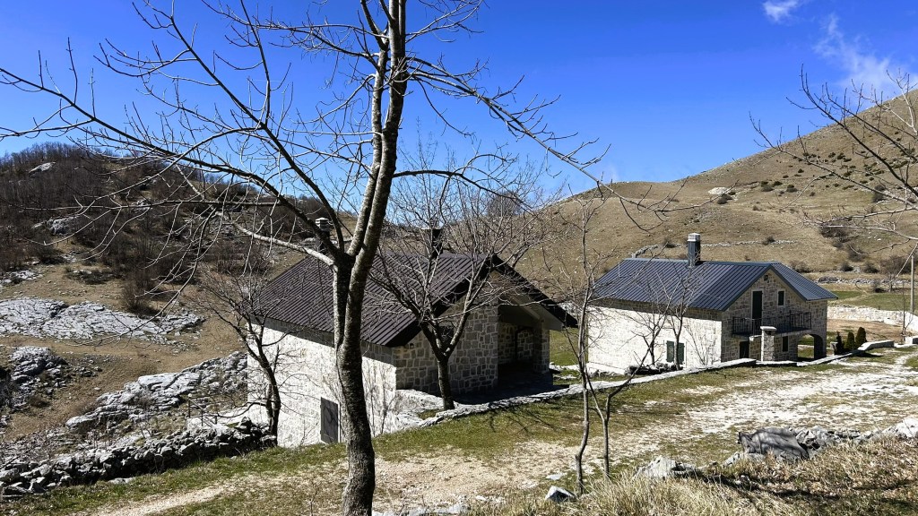 A scenic view of two stone houses set against a backdrop of rolling hills and a clear blue sky, with sparse trees and rocky terrain.