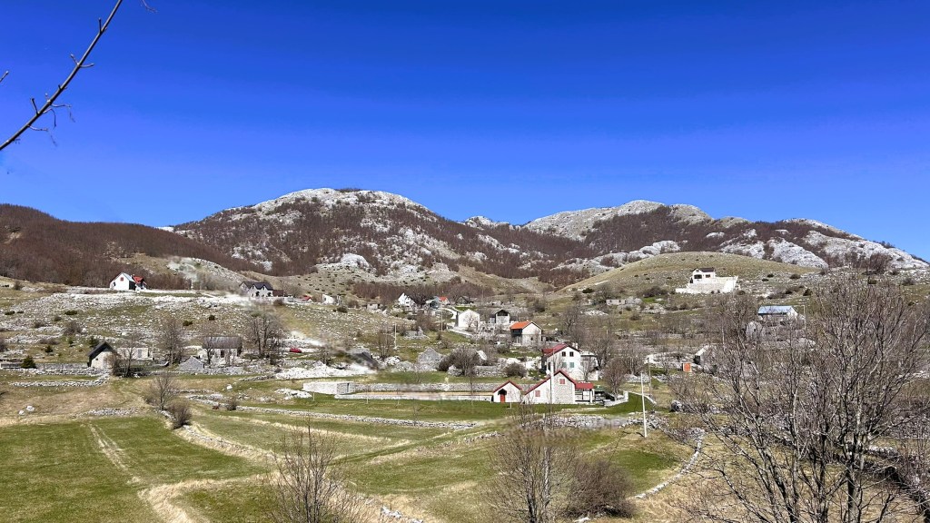 A panoramic view of a picturesque village set against rolling hills and a clear blue sky, featuring various houses with red roofs and green fields.