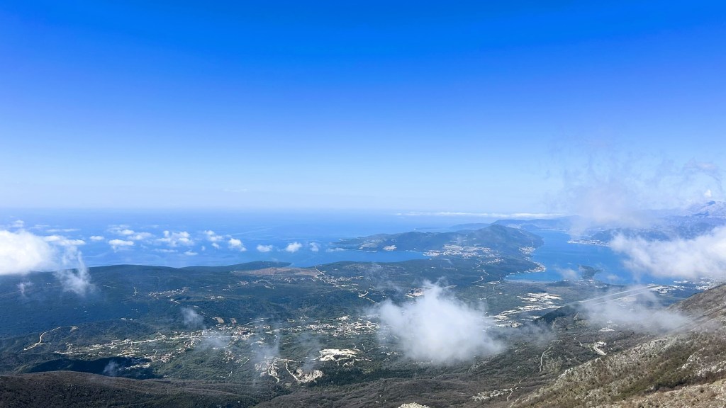 A panoramic view from a mountain top overlooking a lush green landscape, dotted with clouds and leading to a sparkling blue sea under a clear sky.