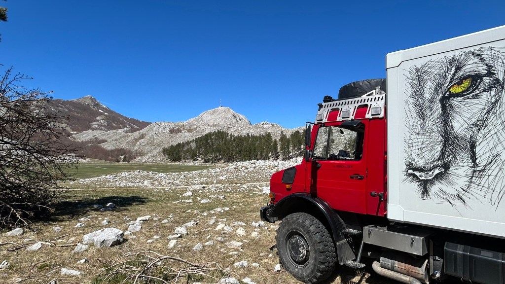 A red off-road truck parked in a mountainous landscape, featuring a detailed lion's face illustration on its side, with clear blue skies and rocky terrain in the background.