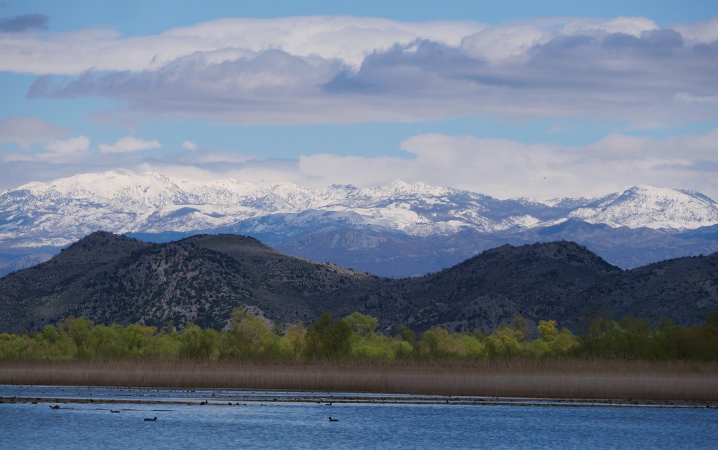 A scenic landscape featuring snow-capped mountains in the background, rolling hills in the midground, and a calm body of water in the foreground, with patches of greenery and clouds in a blue sky.