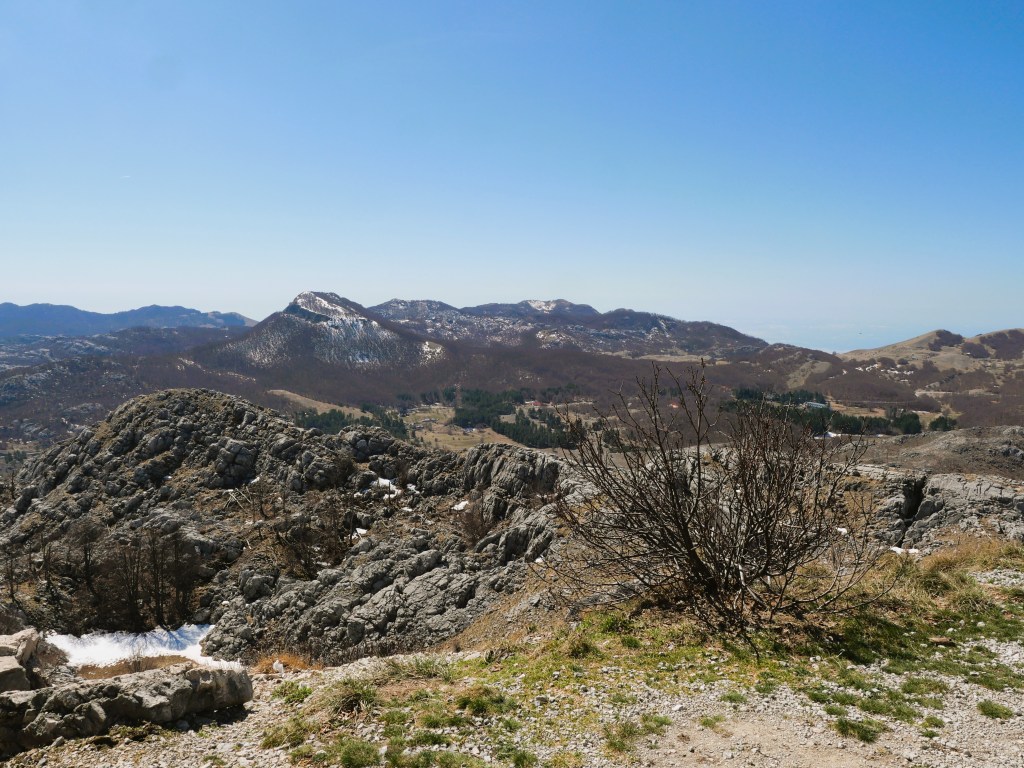 A panoramic view of mountains under a clear blue sky, featuring rocky terrain and sparse vegetation. In the distance, rolling hills can be seen, with some peaks still covered in snow.