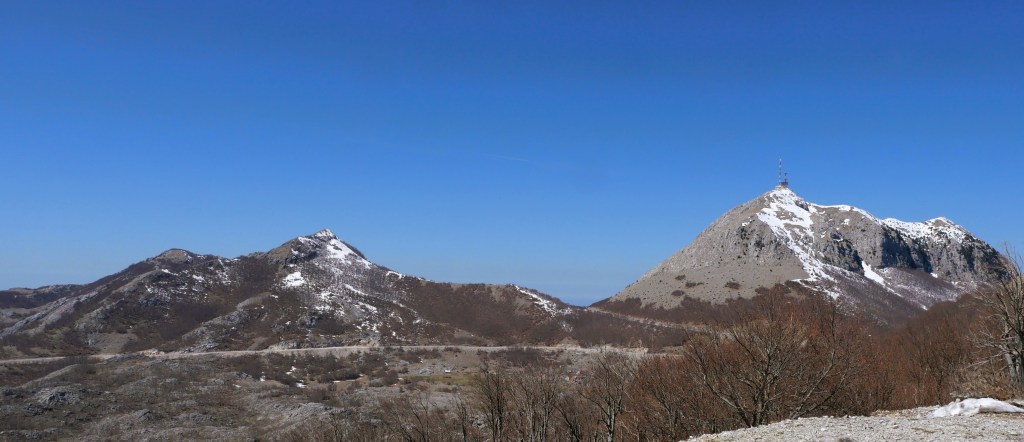 A panoramic view of two snow-capped mountains against a clear blue sky, with a foreground of bare trees and rocky terrain.