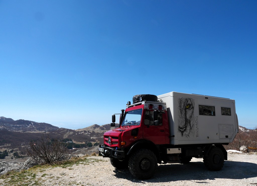 A red off-road vehicle parked on a rocky terrain, featuring a custom camper unit with a lion graphic on its side, set against a clear blue sky and mountainous landscape.